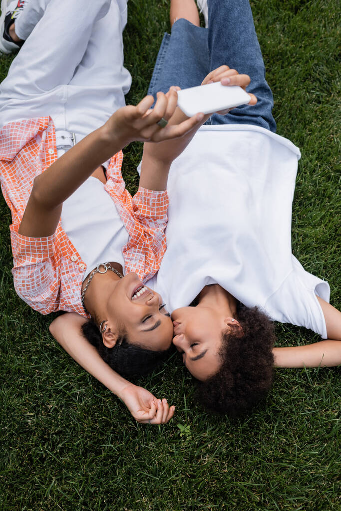 vista superior de la mujer lesbiana afroamericana feliz sosteniendo el teléfono inteligente y tomando selfie con la novia acostada en la hierba  - Foto, Imagen