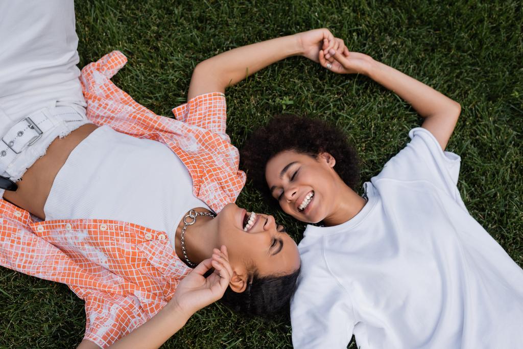 górny widok szczęśliwy afrykański amerykański lesbijki kobiety trzymając się za ręce i leżąc na trawie  - Zdjęcie, obraz