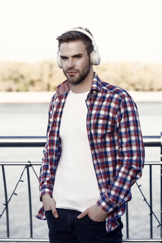 mladý pohledný muž v kostkované košili nosit moderní sluchátka poslech hudby venku, hudba. - Fotografie, Obrázek