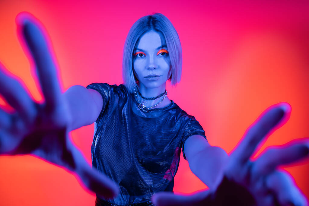 stylish woman with outstretched hands looking at camera in blue neon light on coral and pink background  - Photo, Image