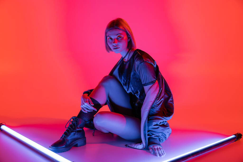 jeune femme à la mode assise près de lampes au néon et regardant la caméra sur fond violet et corail - Photo, image