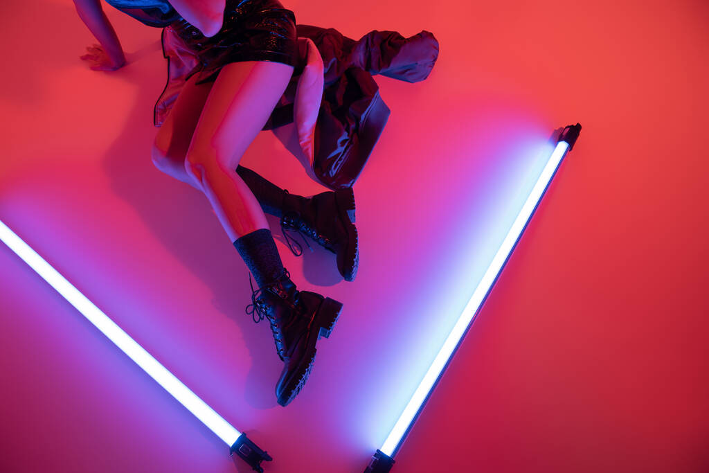 vista dall'alto di donna ritagliata in stivali di pelle nera vicino a lampade al neon viola su sfondo rosso carminio - Foto, immagini