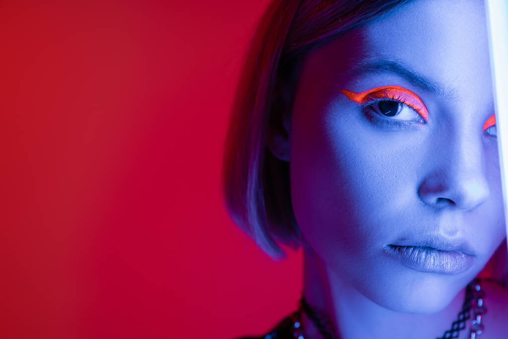 vue recadrée de la femme avec du maquillage au néon regardant la caméra en lumière bleue sur fond rouge carmin - Photo, image
