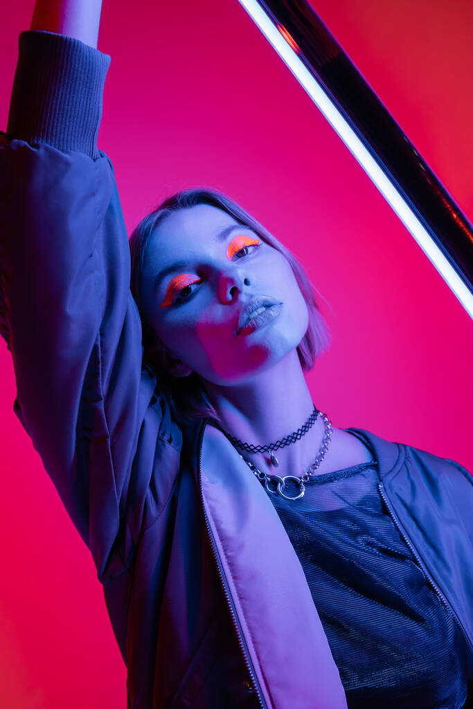 trendy woman with neon makeup and necklaces holding fluorescent lamp on deep pink and coral background - Photo, Image