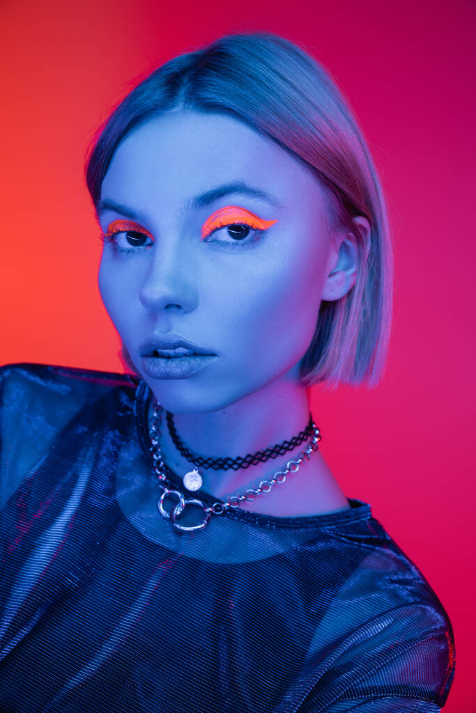 trendi nő nyakláncok és neon smink nézi kamera kék fény korall és rózsaszín háttér - Fotó, kép
