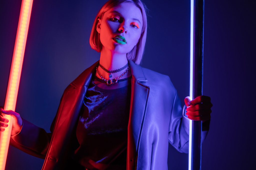 trendy woman in leather jacket holding neon lamps while looking at camera on dark blue background - Photo, Image
