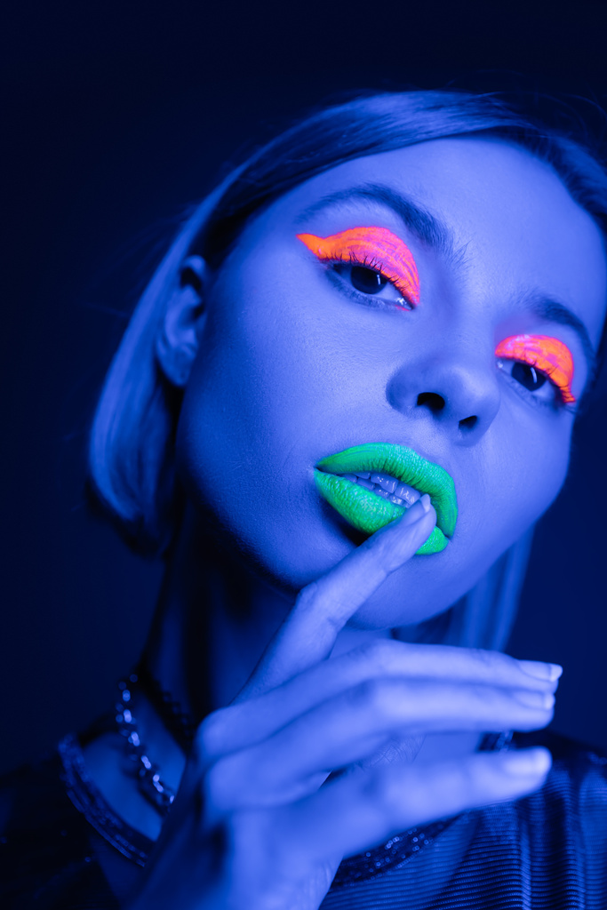 Porträt einer jungen Frau mit buntem Neon-Make-up, die grüne Lippen berührt, isoliert auf dunkelblau - Foto, Bild