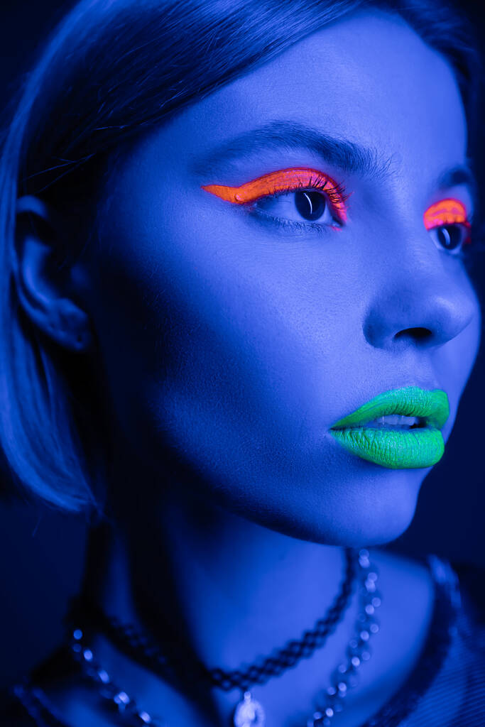 portrait en gros plan de la femme avec un maquillage vibrant regardant loin dans la lumière au néon sur fond bleu foncé - Photo, image