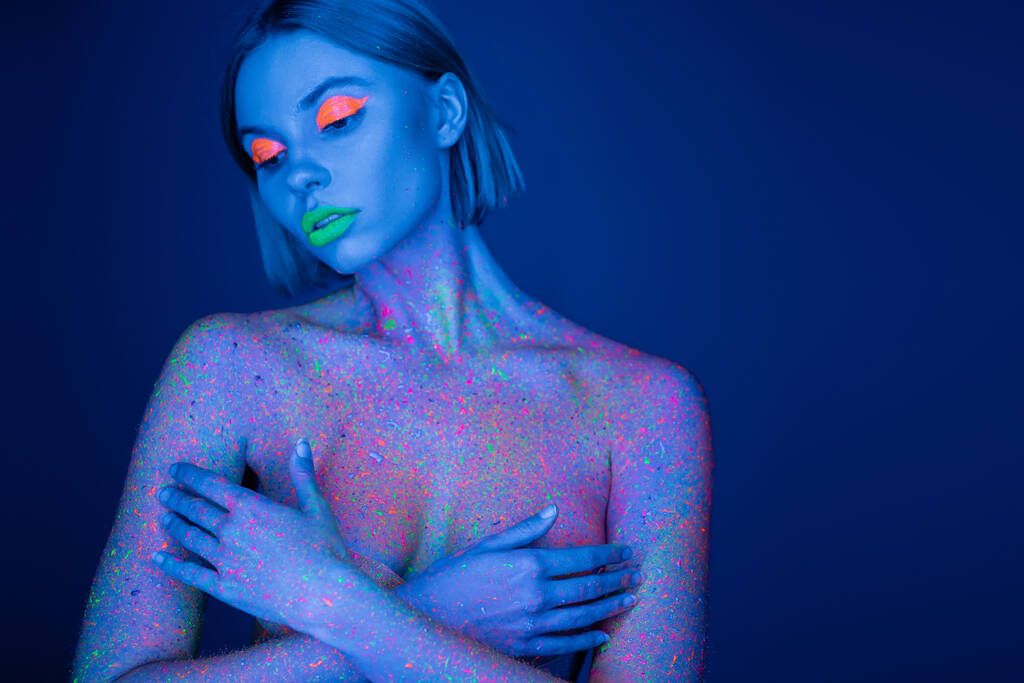 mujer desnuda en maquillaje de neón y salpicaduras de pintura brillante que cubre busto con las manos aisladas en azul oscuro - Foto, Imagen