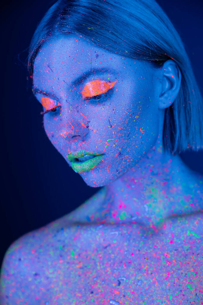 portrait of woman in glowing neon makeup and bright paint splashes isolated on dark blue - Photo, Image