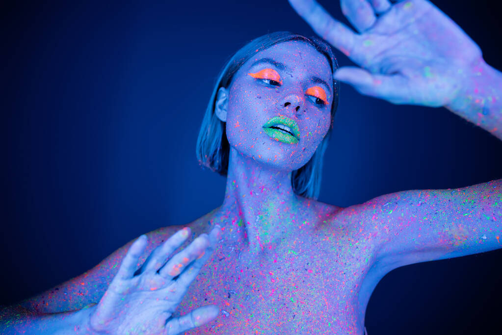 meztelen és érzéki nő neon smink és színes test pózol sötétkék háttér - Fotó, kép