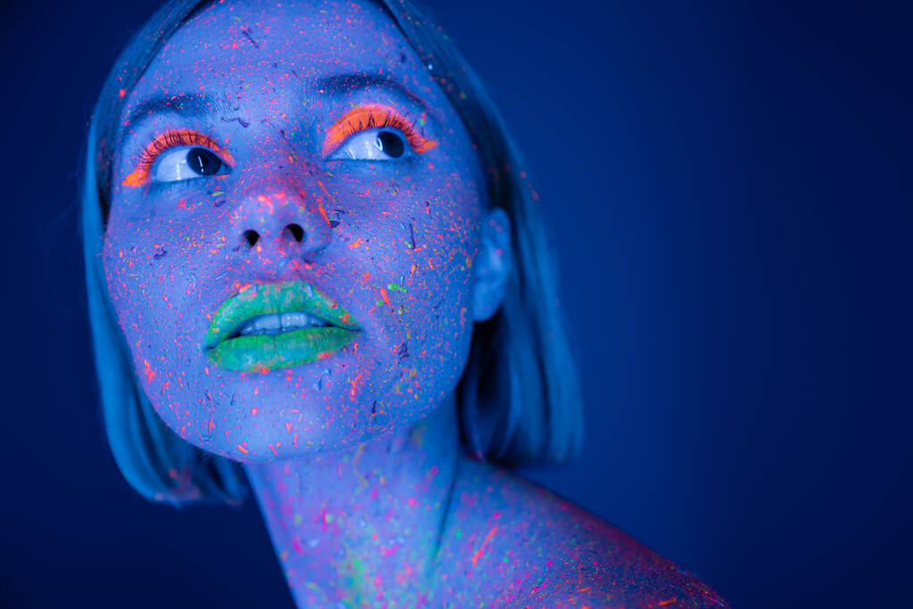 Porträt einer jungen Frau mit Neon-Make-up und lebhafter Farbe im Gesicht, die isoliert auf dunkelblau wegschaut - Foto, Bild