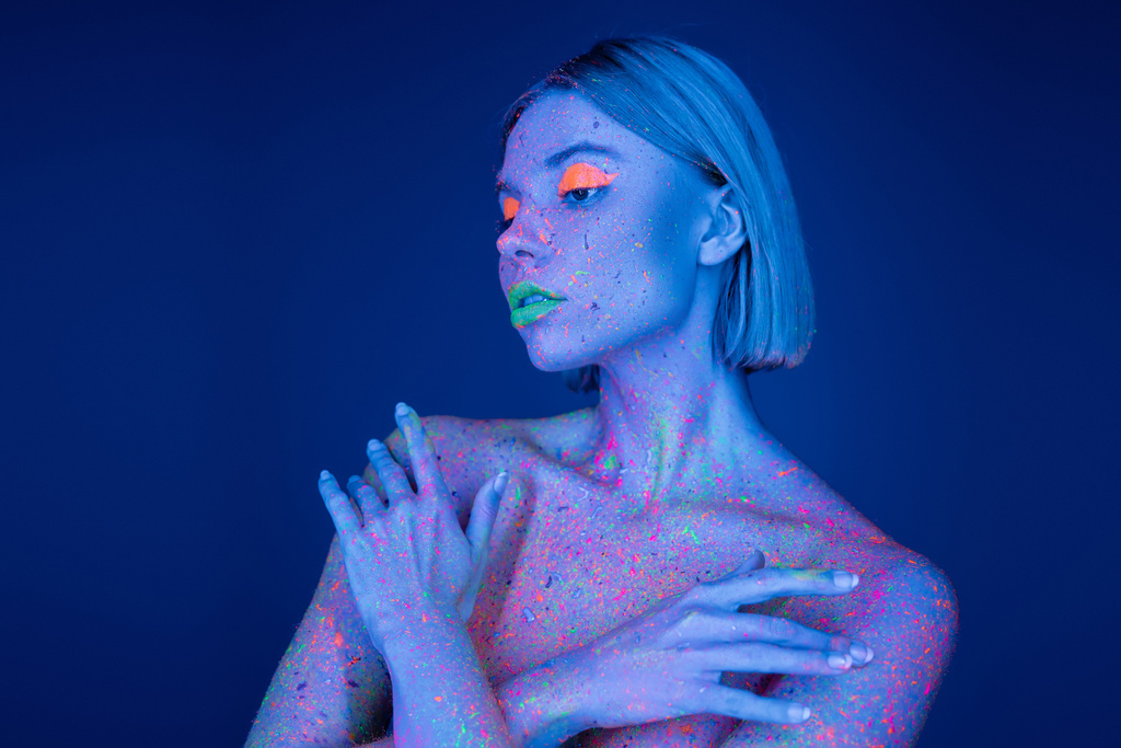 обнаженная женщина в неоновом макияже и светящиеся краски брызги на теле позируют со скрещенными руками изолированы на синий - Фото, изображение