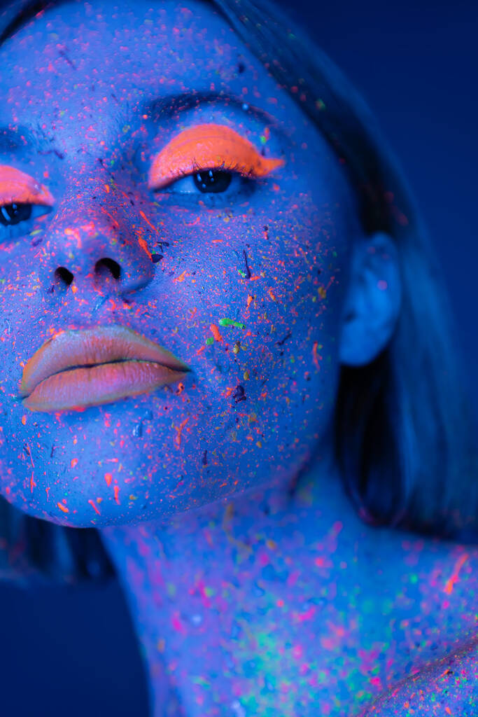 Nahaufnahme Porträt einer Frau in leuchtendem Neon-Make-up und heller Farbe isoliert auf dunkelblau - Foto, Bild