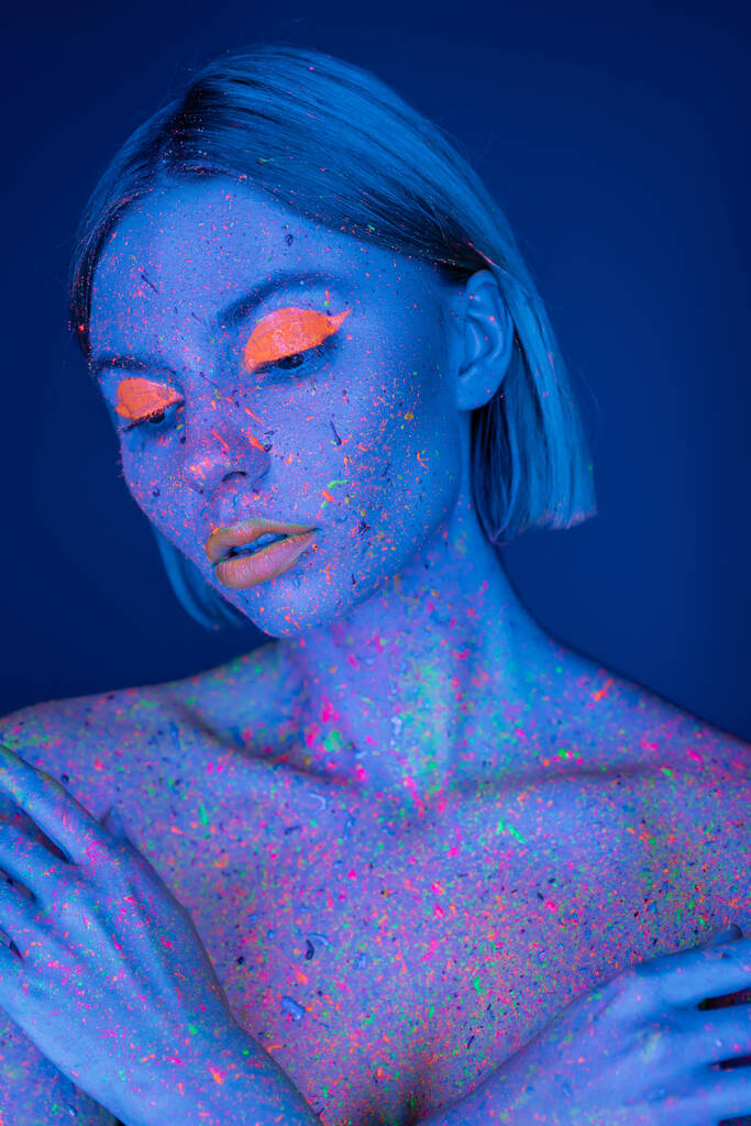mujer desnuda en maquillaje de neón brillante y salpicaduras de pintura fluorescente posando aislado en azul oscuro - Foto, Imagen