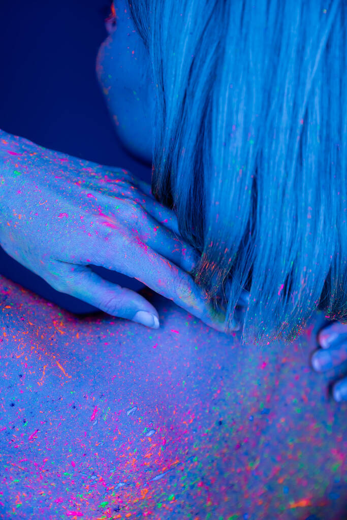 μερική άποψη της γυναίκας αγγίζοντας το σώμα χρωματισμένο με φωτεινό νέον χρώμα που απομονώνονται σε σκούρο μπλε - Φωτογραφία, εικόνα