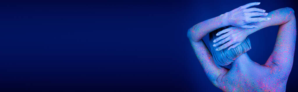 vista posterior de la mujer de color con pintura de neón posando con las manos detrás de la cabeza aislada en azul oscuro, bandera - Foto, Imagen