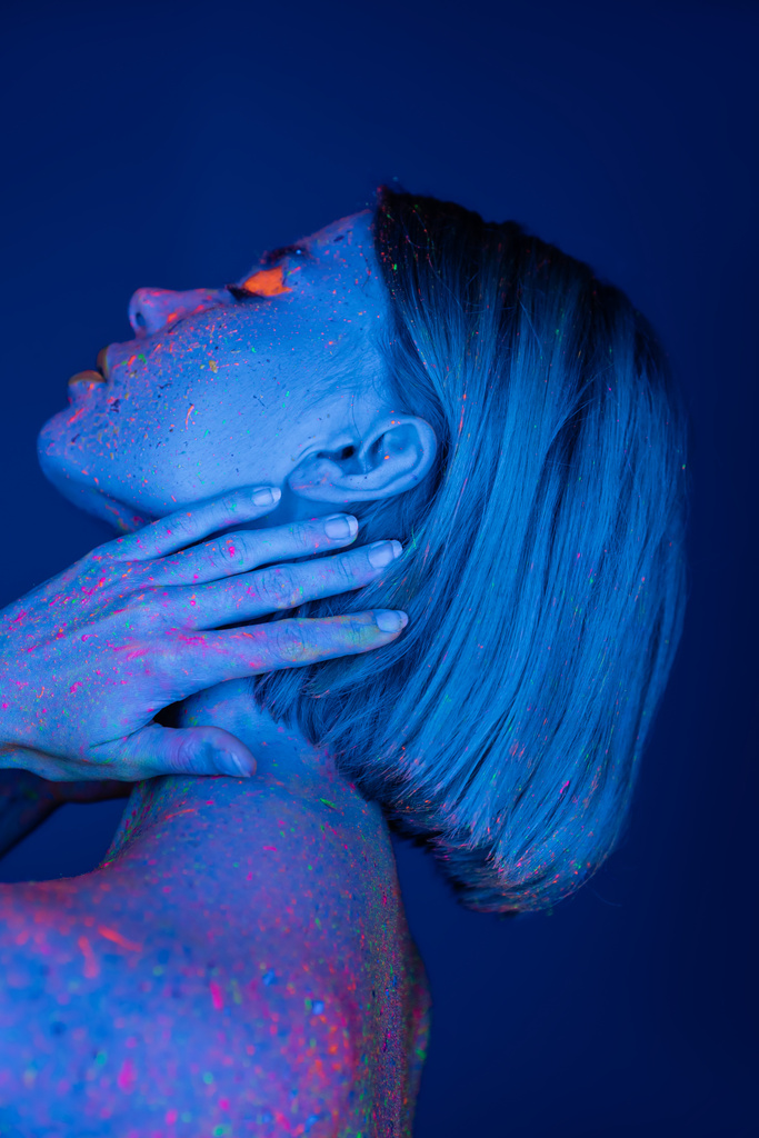 vista lateral da mulher em brilhante neon corpo pintura tocando pescoço isolado no azul escuro - Foto, Imagem