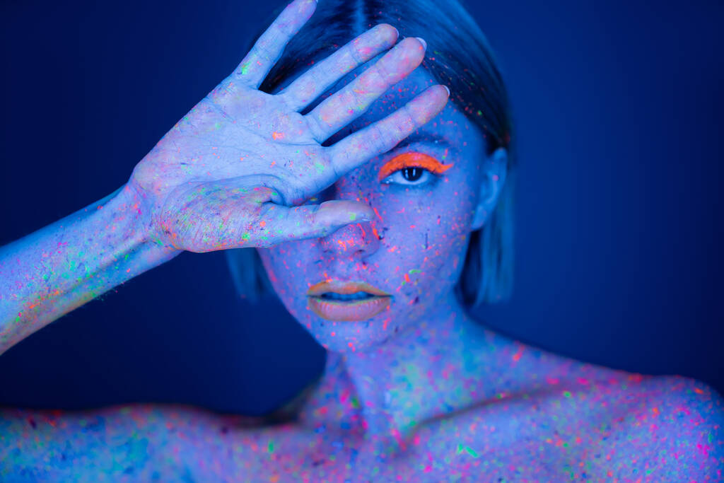 mujer en maquillaje de neón y pintura corporal brillante que oscurece la cara con la mano mientras mira a la cámara aislada en azul oscuro - Foto, Imagen
