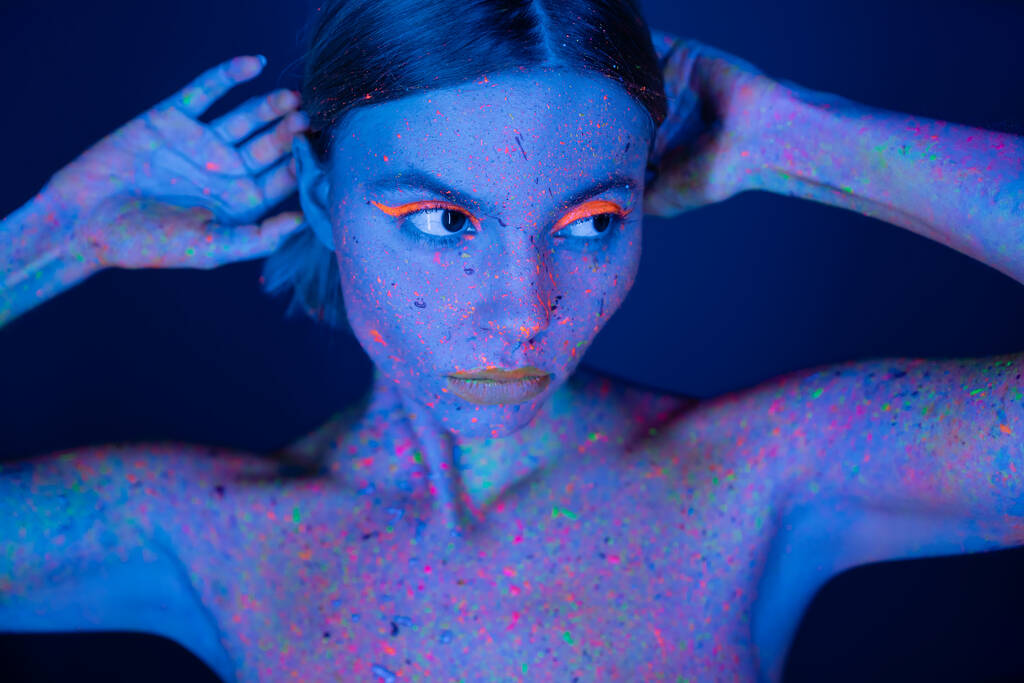 junge Frau in leuchtendem Neon-Make-up und farbenfroher Körperfarbe, die isoliert auf dunkelblauem Grund wegschaut - Foto, Bild