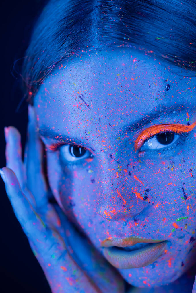 close-up portret van vrouw met neon make-up en kleurrijke verf op gezicht op zoek naar camera geïsoleerd op donkerblauw - Foto, afbeelding