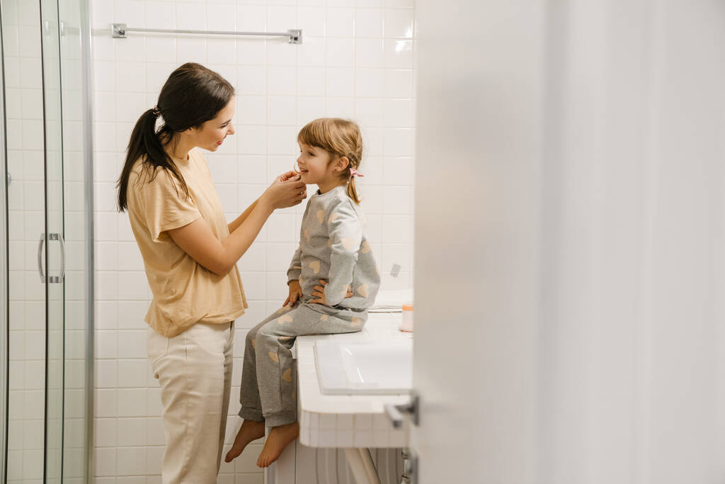 Valkoinen nainen hymyilee puhuessaan tyttärensä kanssa kylpyhuoneessa kotona - Valokuva, kuva