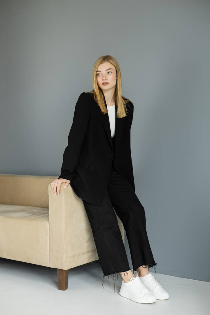 Полная длина красивая блондинка в куртке, стоящая рядом с диваном дома  - Фото, изображение