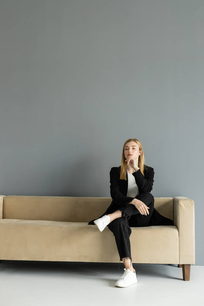 Donna pensierosa in giacca seduta sul divano beige a casa  - Foto, immagini