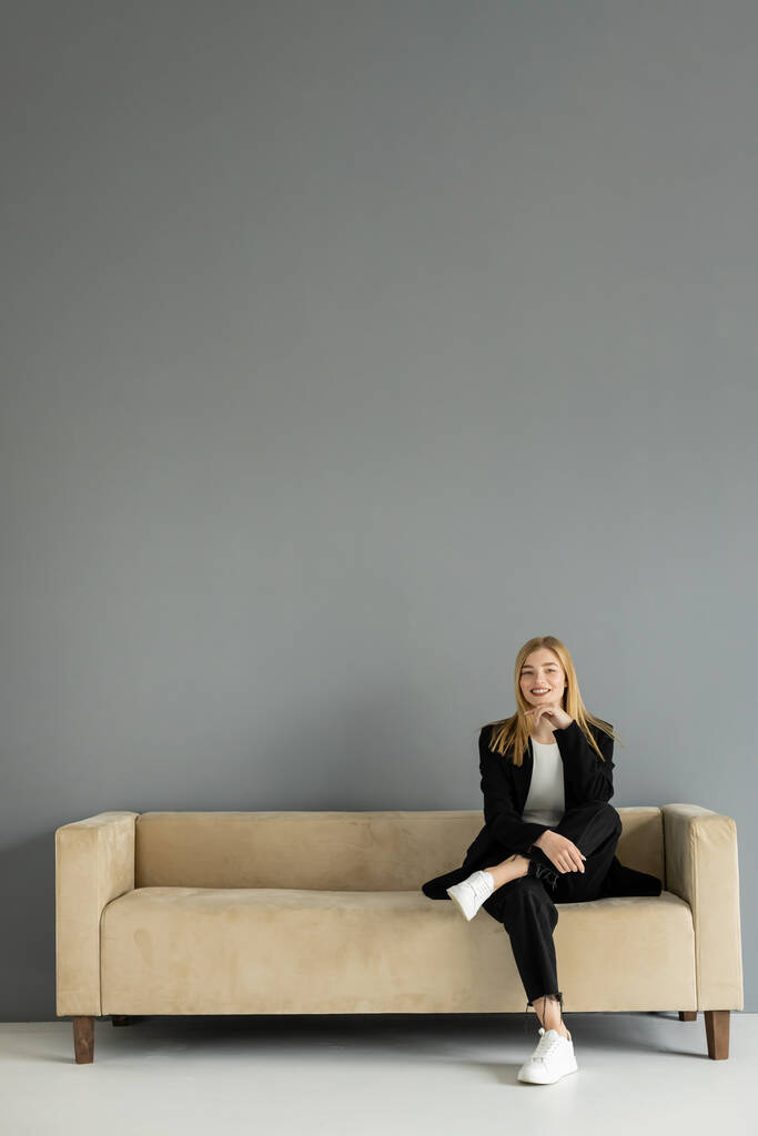 Fröhliche blonde Frau blickt in die Kamera, während sie auf einer Couch in der Nähe einer grauen Wand sitzt  - Foto, Bild