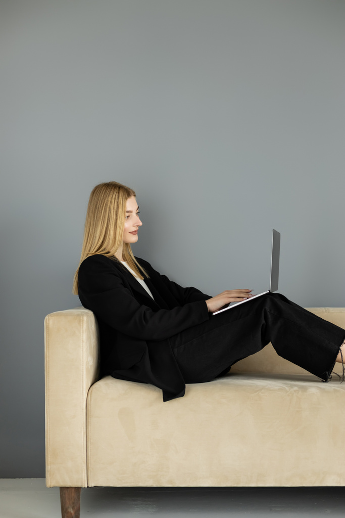Treinador sorridente na jaqueta usando laptop no sofá perto da parede cinza  - Foto, Imagem