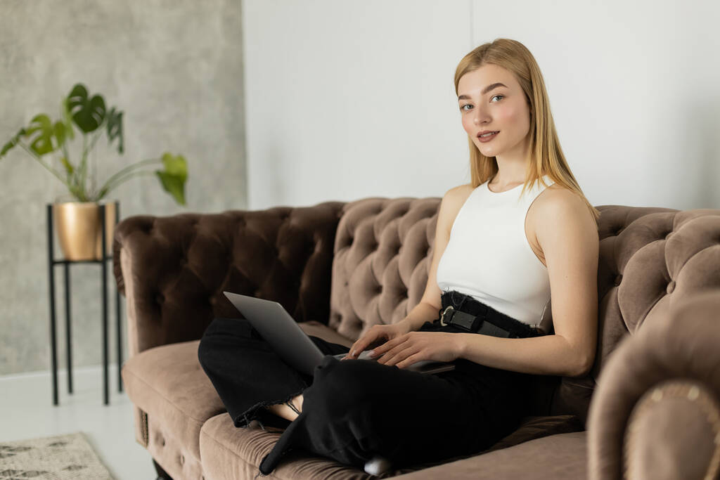 Femme blonde souriante en haut en utilisant un ordinateur portable et en regardant la caméra sur le canapé  - Photo, image