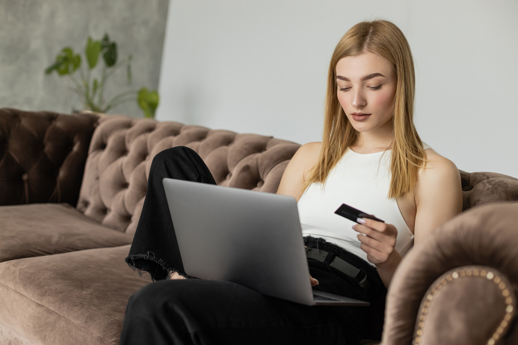 Blonde woman in top holding credit card and using laptop on comfortable couch  - Photo, Image