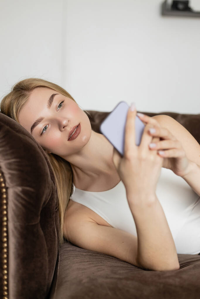 Kaunis nuori nainen katselee hämärtynyt matkapuhelin ruskea sohva kotona  - Valokuva, kuva