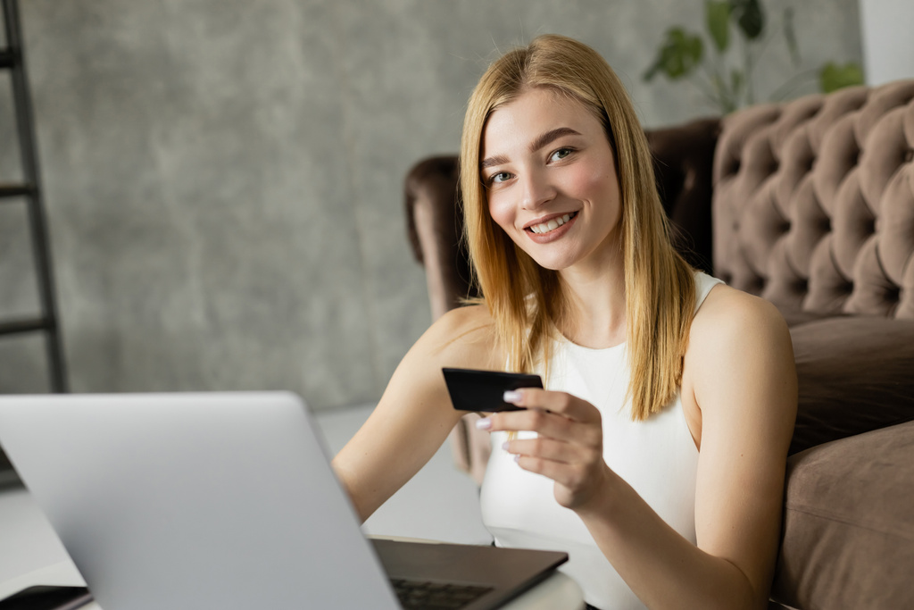 Mujer rubia sonriendo a la cámara mientras sostiene la tarjeta de crédito cerca de la computadora portátil en la sala de estar  - Foto, imagen