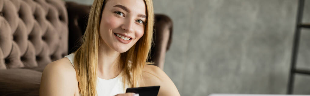 Ξανθιά γυναίκα χαμογελά στην κάμερα, ενώ κρατώντας πιστωτική κάρτα στο σπίτι, πανό  - Φωτογραφία, εικόνα