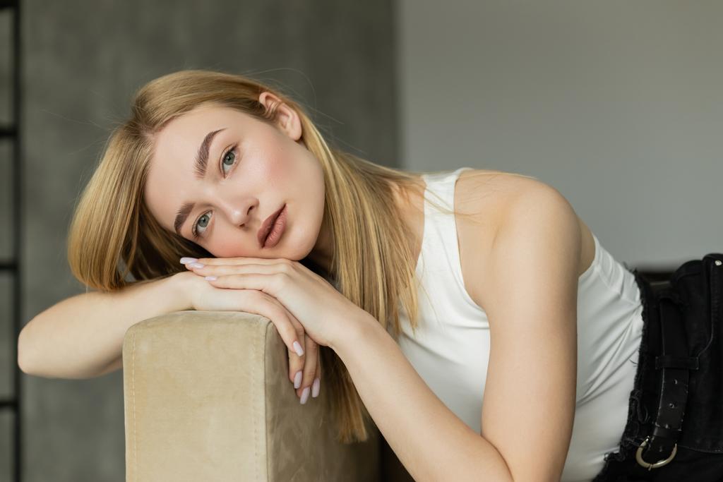 Young blonde woman in top looking at camera while relaxing on couch  - Photo, Image
