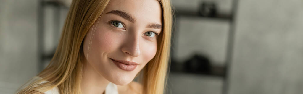 Mujer rubia joven sonriendo a la cámara en la sala de estar borrosa, pancarta  - Foto, Imagen