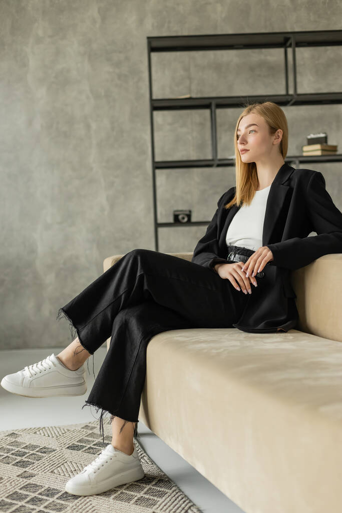 full length of stylish and pensive woman in black clothes and white sneakers looking away on couch in loft style living room - Photo, Image