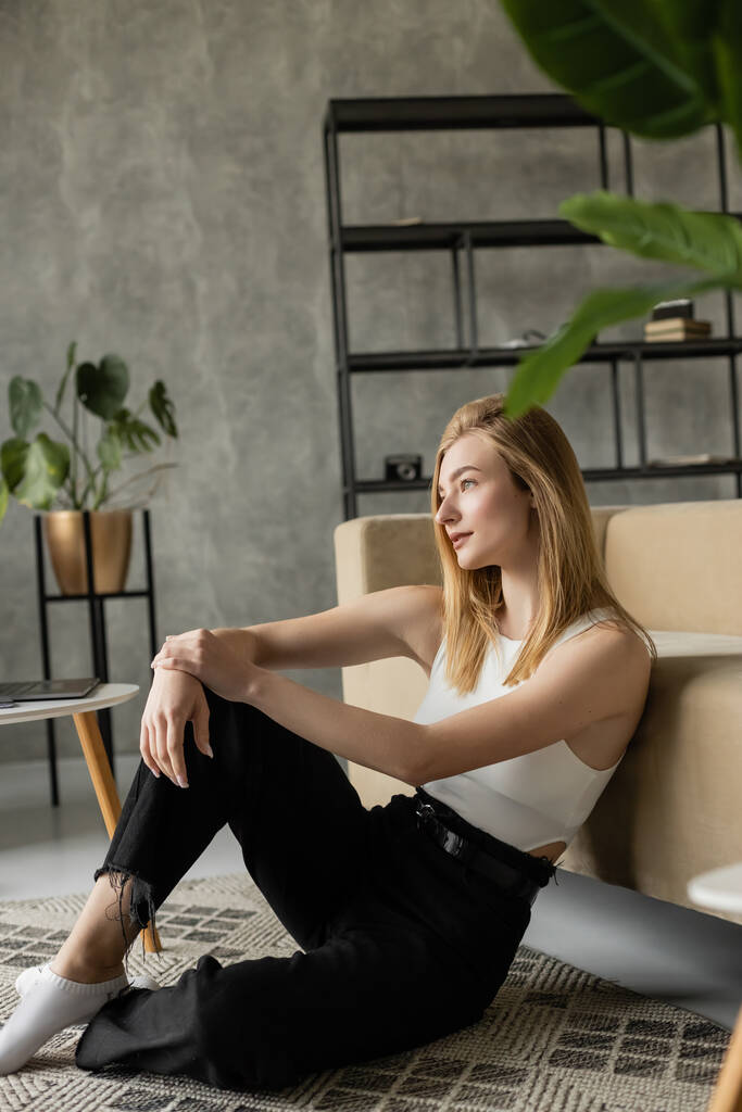 mulher loira pensativa em jeans pretos sentado no chão perto do sofá e olhando para longe no apartamento estilo loft - Foto, Imagem