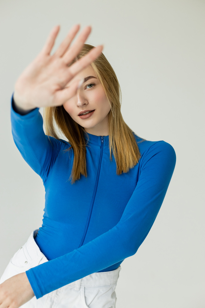 trendikäs nainen sininen pitkähihainen paita seisoo ojennettu käsi hämärtynyt etualalla eristetty harmaa - Valokuva, kuva