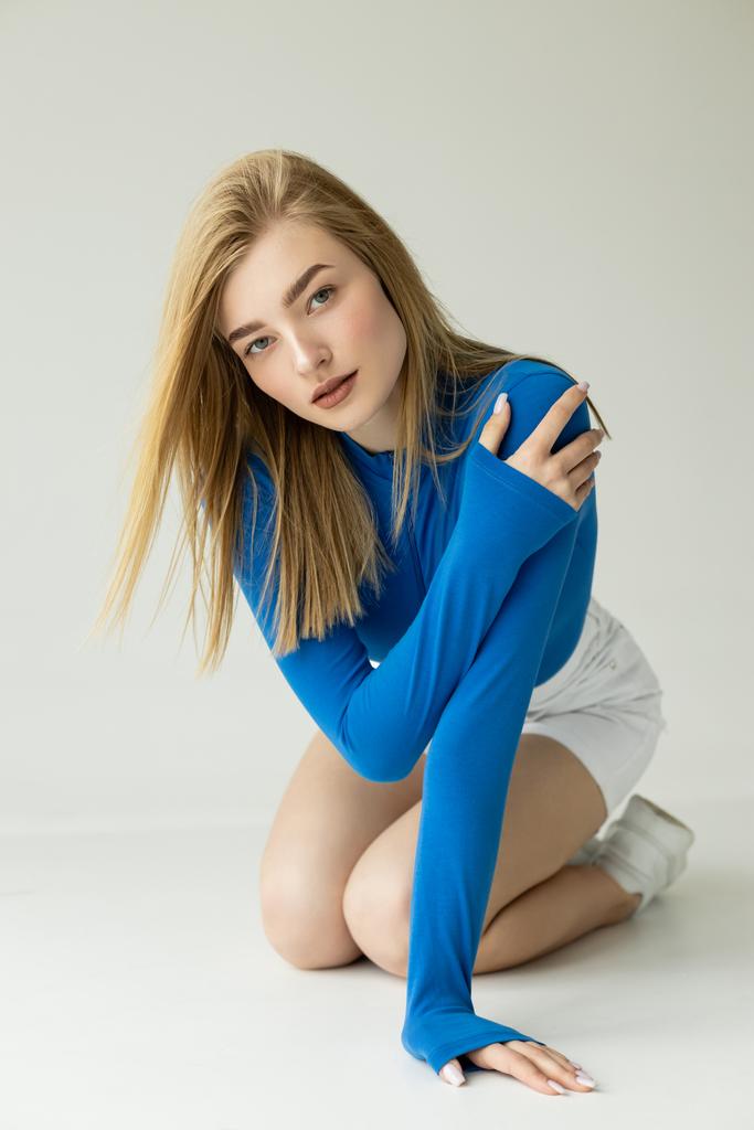 sensual blonde woman in blue long sleeve shirt looking at camera while posing on knees on grey background  - Photo, Image