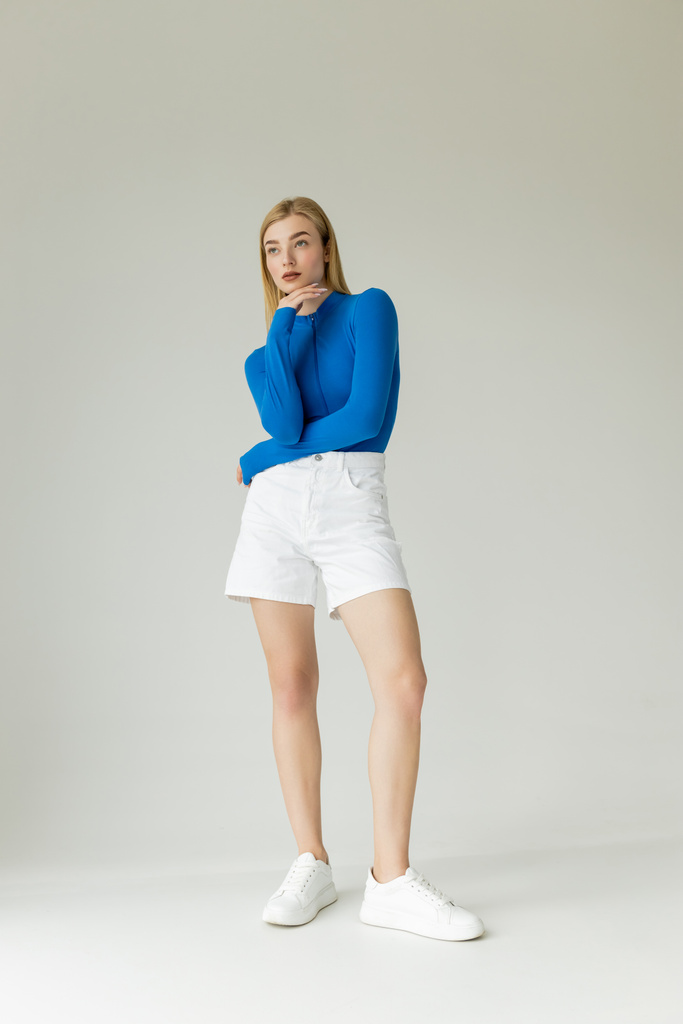Полная длина стройная женщина в голубой рубашке с длинным рукавом и белые шорты с кроссовками глядя в сторону на сером фоне  - Фото, изображение