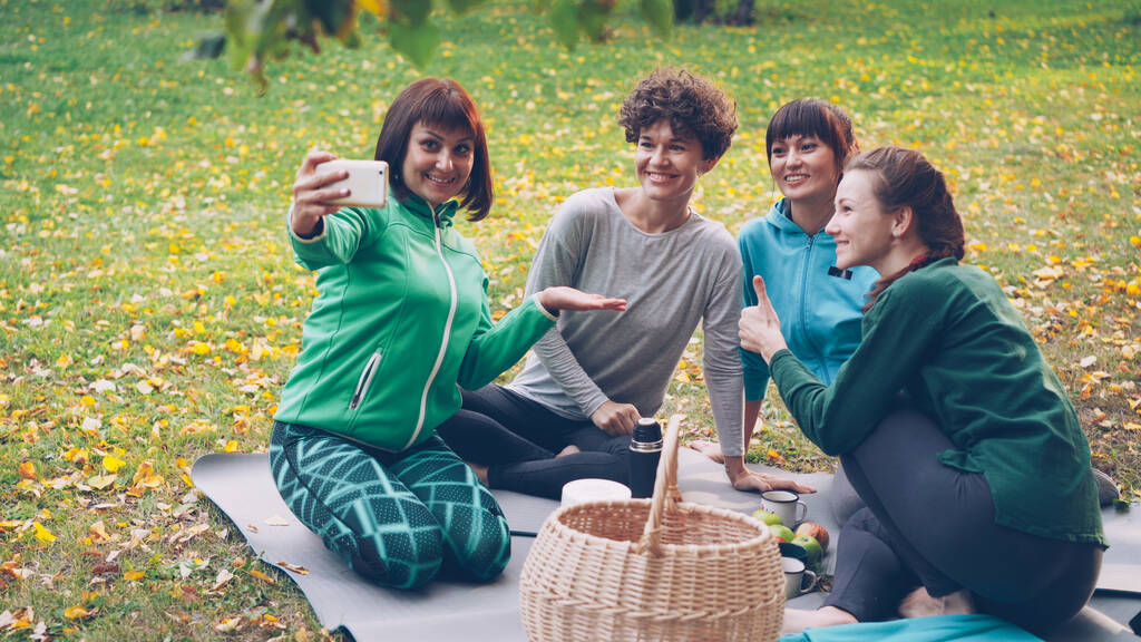 Досить молоді жінки беруть селфі зі смартфоном під час пікніка в парку, сидячи на траві, позуючи і сміючись розважаючись. Природа, дружба та концепція сучасних технологій
. - Фото, зображення