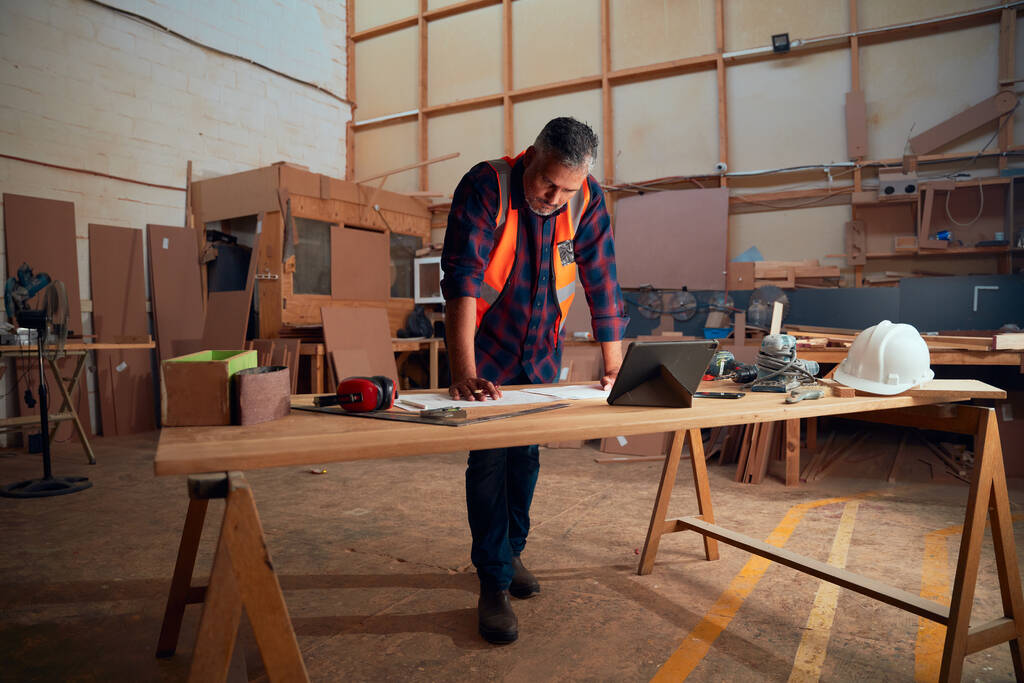 Multiracial hombre adulto medio trabajando junto a la tableta digital y herramientas en la mesa de madera en la fábrica de carpintería - Foto, Imagen