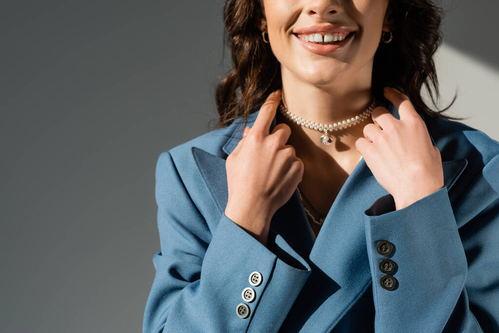 vue recadrée d'une femme souriante en collier de perles et blazer bleu sur fond gris - Photo, image
