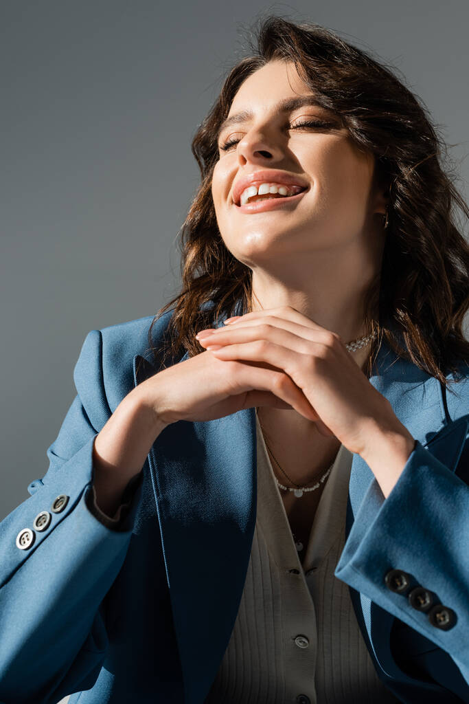 excitée femme brune en blazer bleu et à la mode riant avec les yeux fermés isolé sur gris - Photo, image