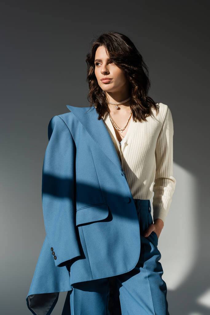 модная брюнетка с голубым блейзером, стоящая с рукой в кармане и глядя в сторону на сером фоне - Фото, изображение