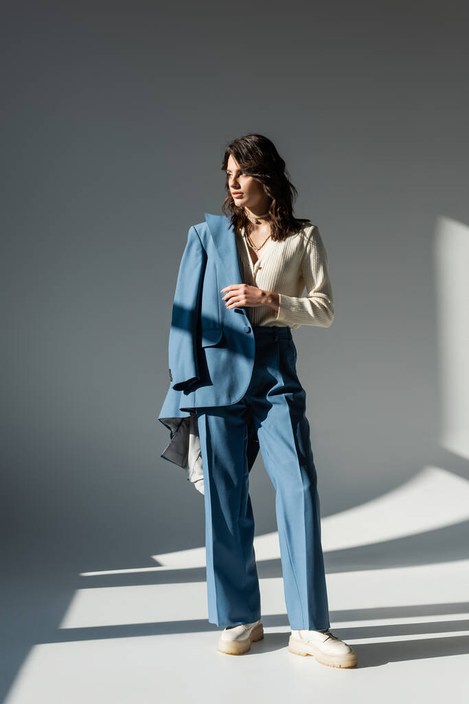 полная длина модной женщины в белых сапогах, стоящей в синей куртке на сером фоне с освещением и тенями - Фото, изображение