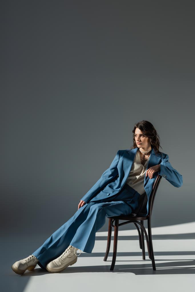 teljes hossza elegáns nő kék öltöny és csizma néz félre a szék szürke háttér világítás és árnyékok - Fotó, kép