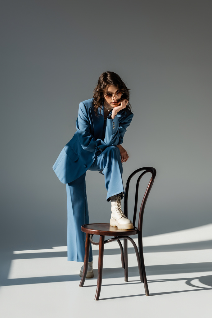 full length of brunette woman in blue suit and trendy sunglasses stepping on chair on grey background with lighting - Photo, Image